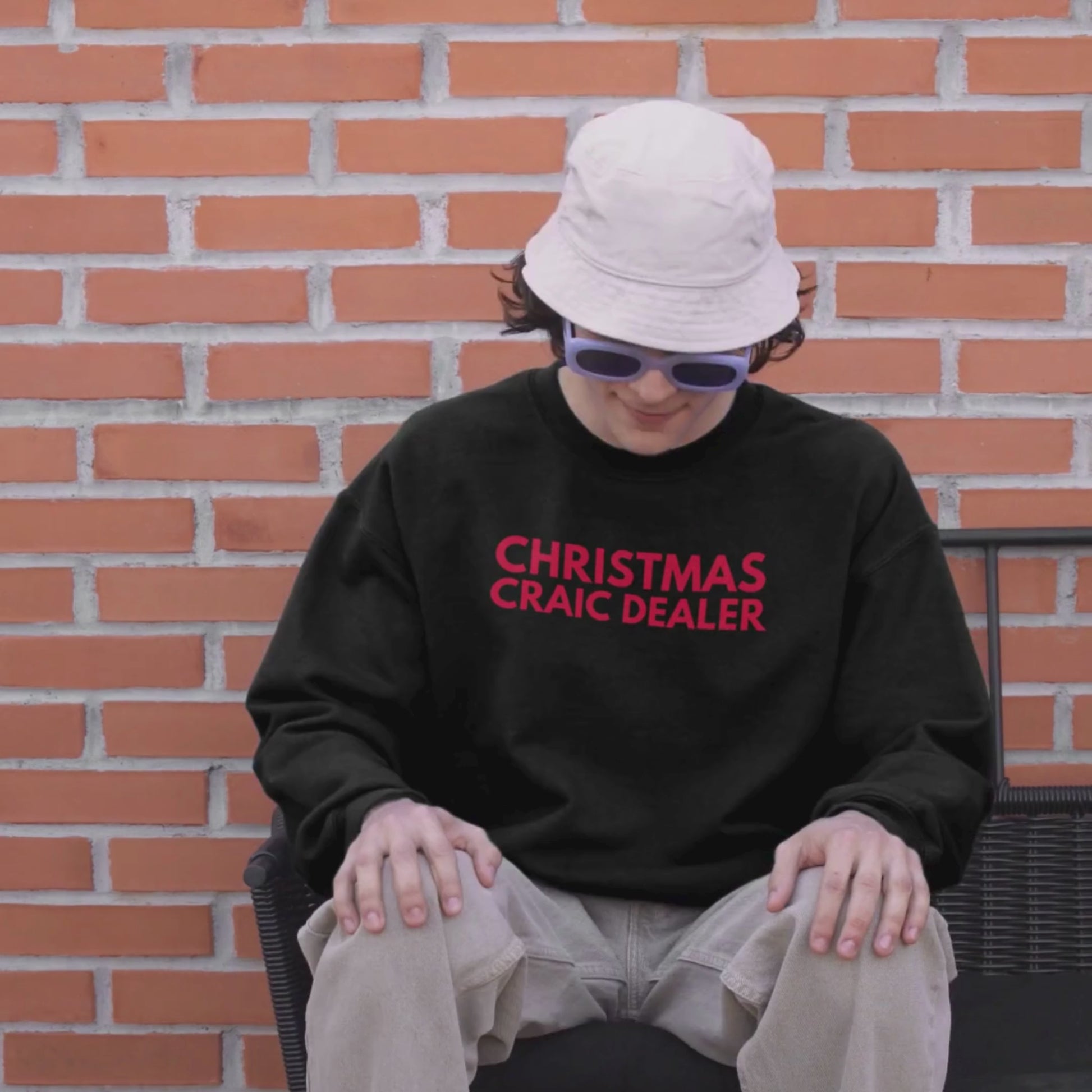 Person wearing a beige bucket hat and black sweatshirt with red text 'CHRISTMAS CRAIC DEALER' sitting in front of a red brick wall.