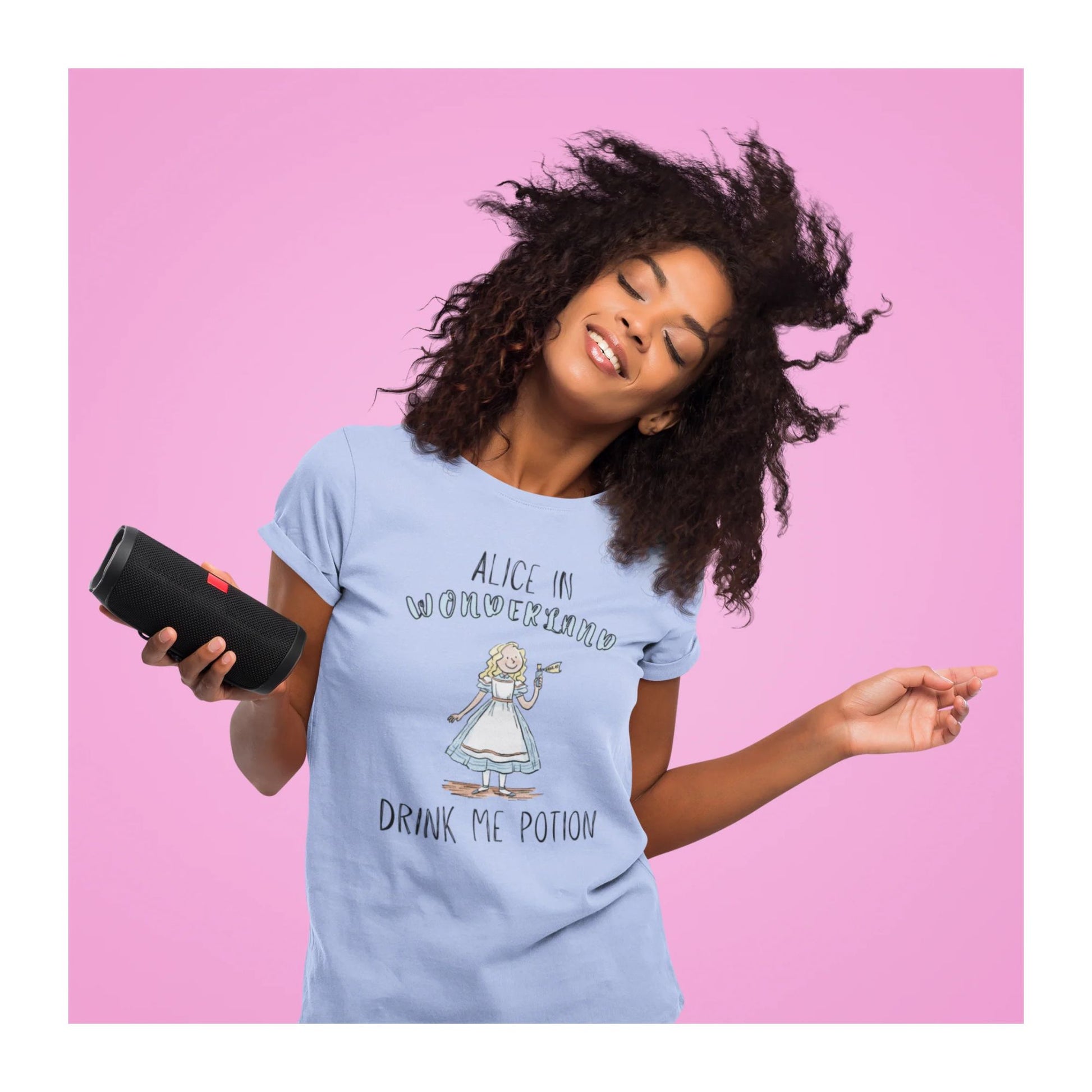 Woman wearing a light blue t-shirt with 'Alice in Wonderland' design, holding a black speaker against a pink background.