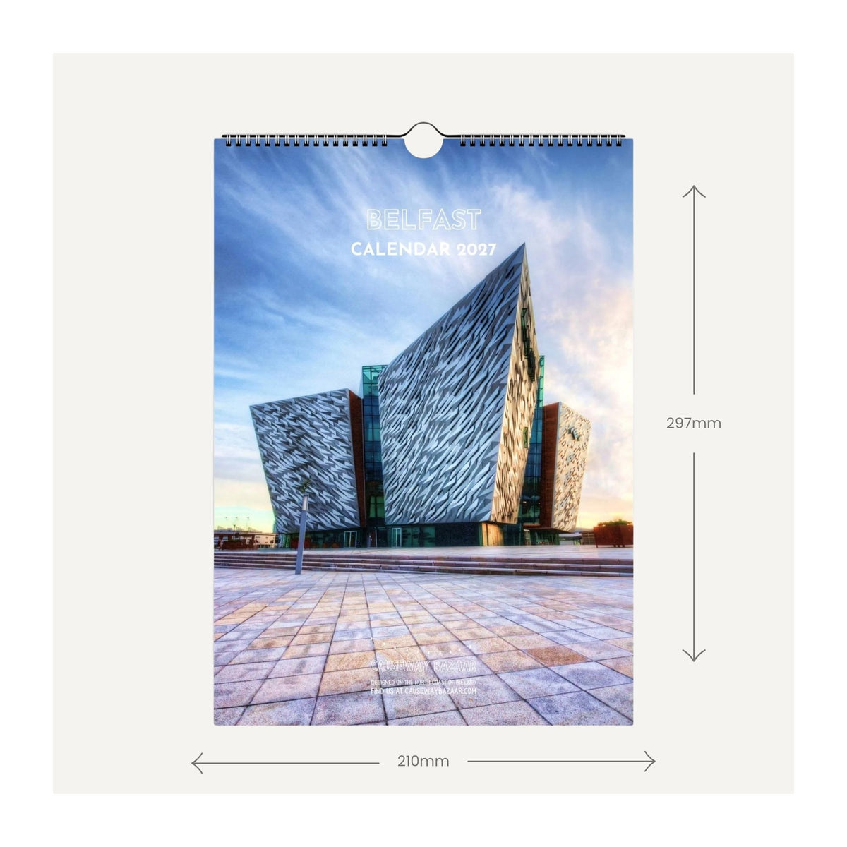 A hanging wall calendar cover shown with dimension arrows and labels indicating 210mm width and 297mm height, on a light beige background. The calendar features a photograph of the Belfast skyline, with the words 'BELFAST CALENDAR 2027' printed near the top.