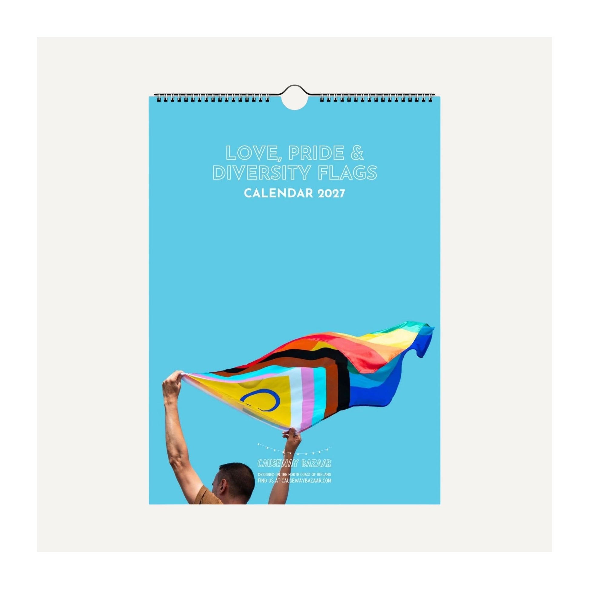 A 2027 wall calendar titled 'LOVE, PRIDE & DIVERSITY FLAGS CALENDAR 2027' featuring a bright blue cover with an illustration of a person holding a rainbow pride flag. The calendar is shown hanging against a light beige background.