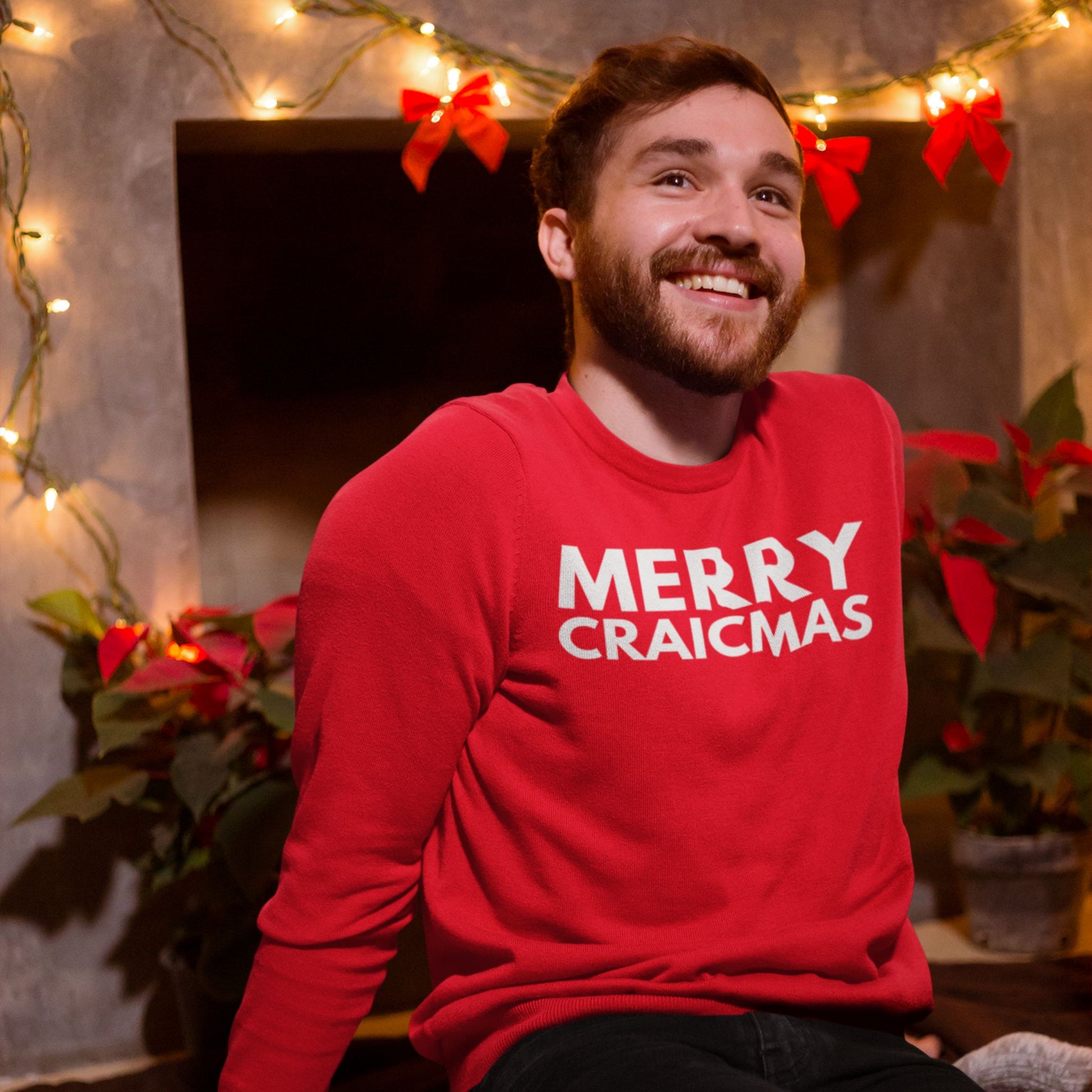 Man wearing a red sweater with 'MERRY CRAICMAS' text in light pink, surrounded by Christmas decorations.