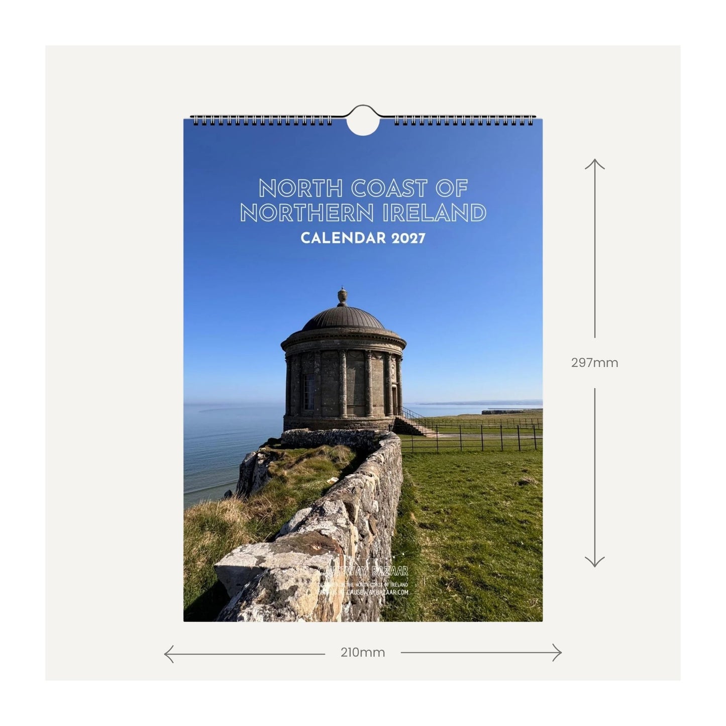 A wall calendar cover shown as a product mockup with dimension arrows and labels. The cover features a photograph of Mussenden Temple at Downhill, situated on a grassy coast under a blue sky, with the text 'NORTH COAST OF NORTHERN IRELAND CALENDAR 2027'.