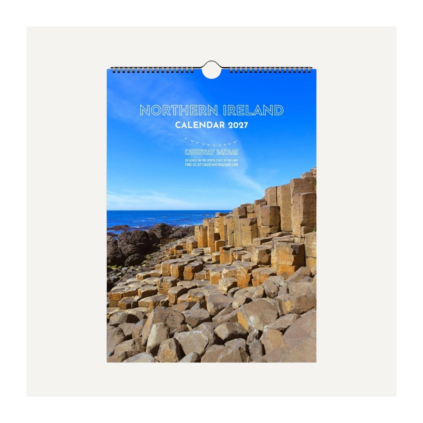 A hanging wall calendar for Northern Ireland 2027, featuring a scenic photograph of the Giant's Causeway rock formations under a bright blue sky. The calendar is shown against a light grey background.