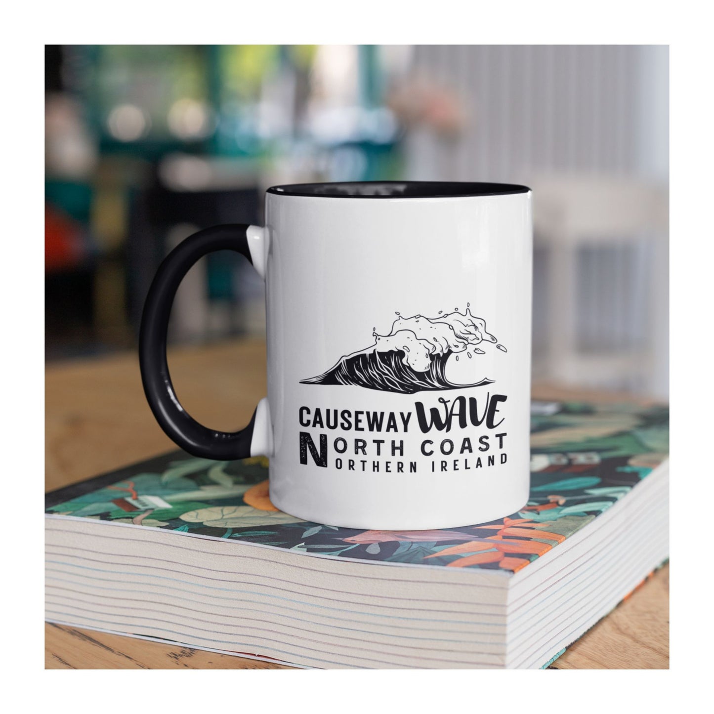 White mug with black handle featuring a wave design and Causeway Wave North Coast Northern Ireland text on a white background placed on a book