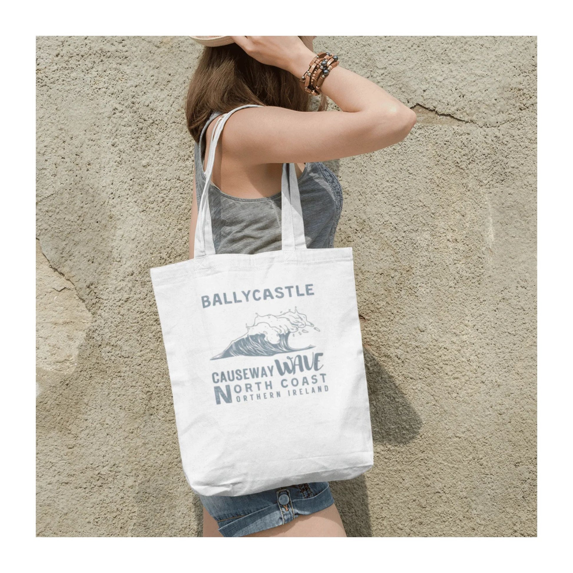 Person holding a tote bag with 'Ballycastle Causeway Way North Coast Northern Ireland' text against a textured wall.
