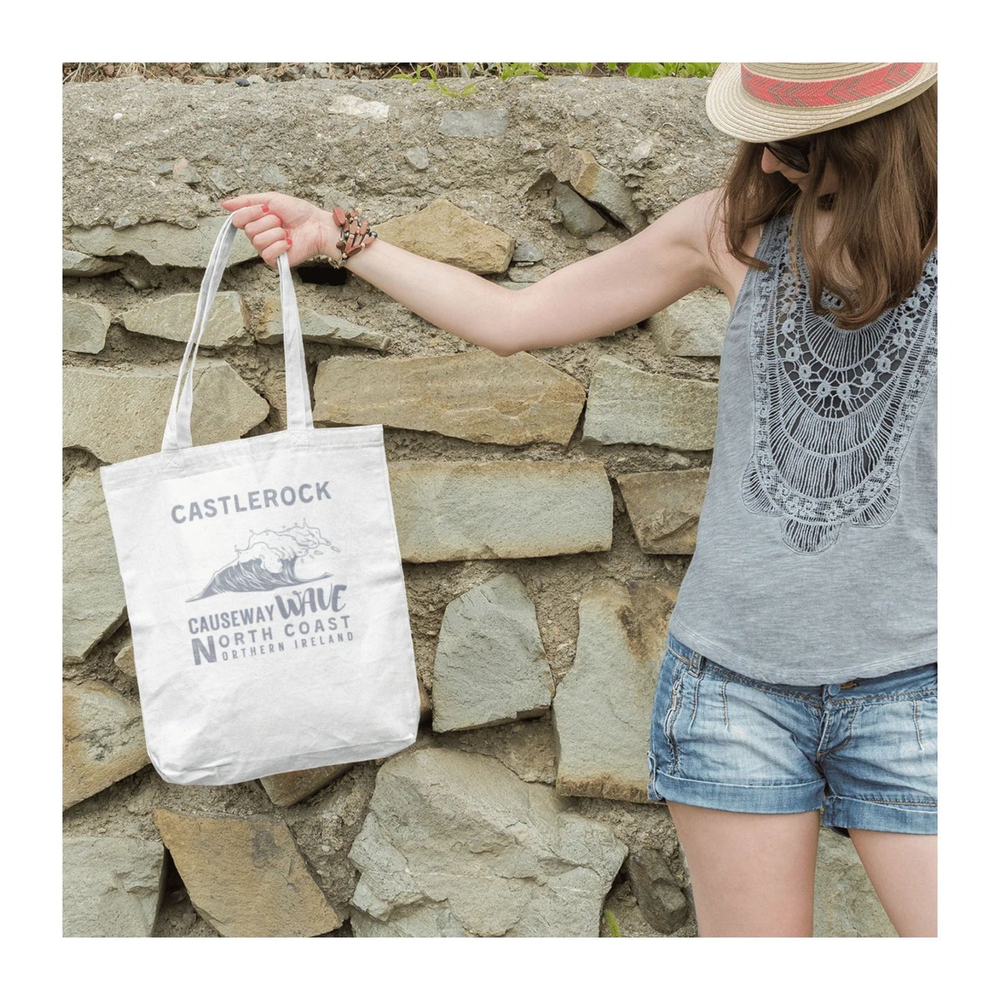 Person holding a white tote bag with 'Castlerock Causeway Wave North Coast Northern ireland' branding against a stone wall.