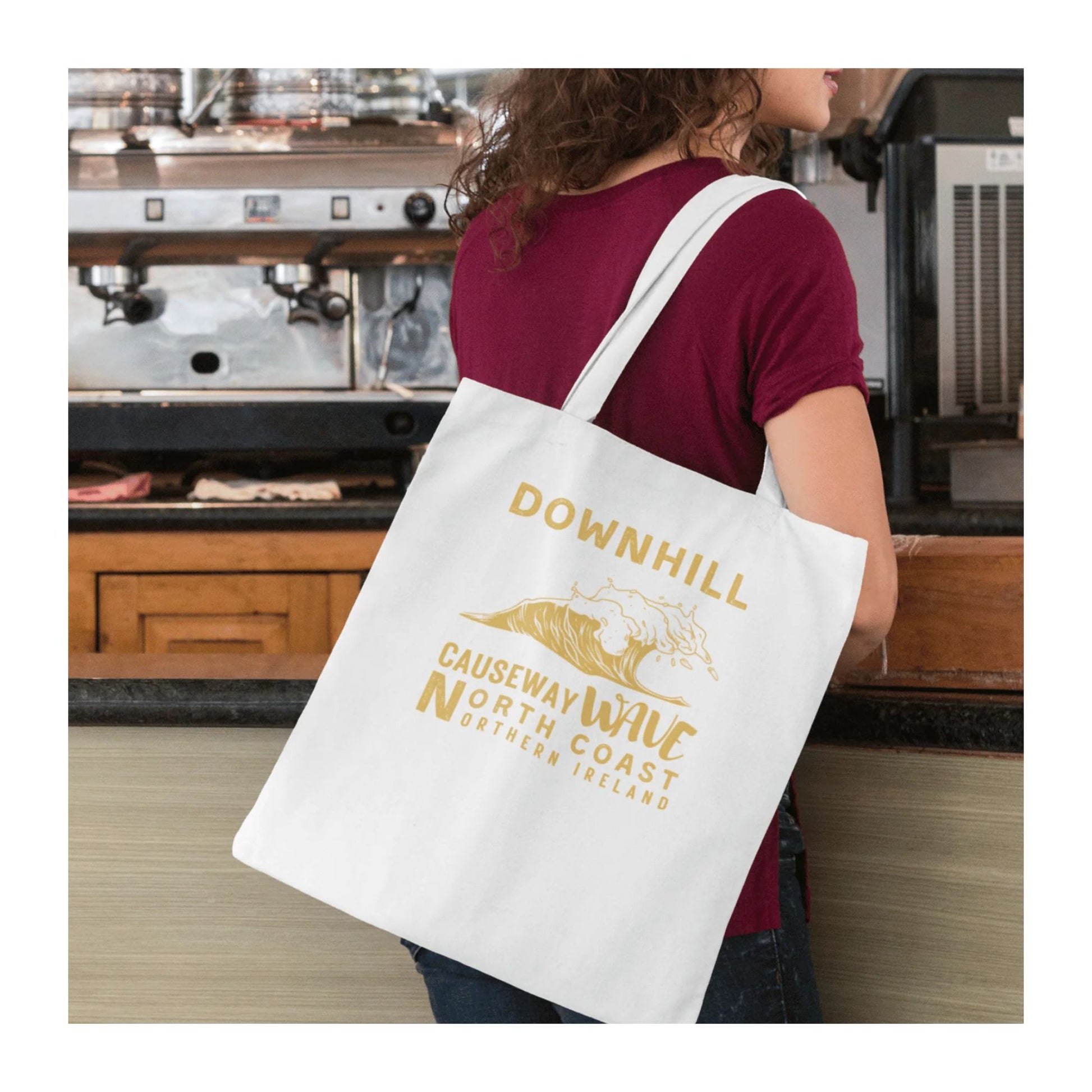 Person holding a white tote bag with yellow text 'Downhill Causeway Wave North Coast Northern Ireland' and wave graphics in a coffee shop.