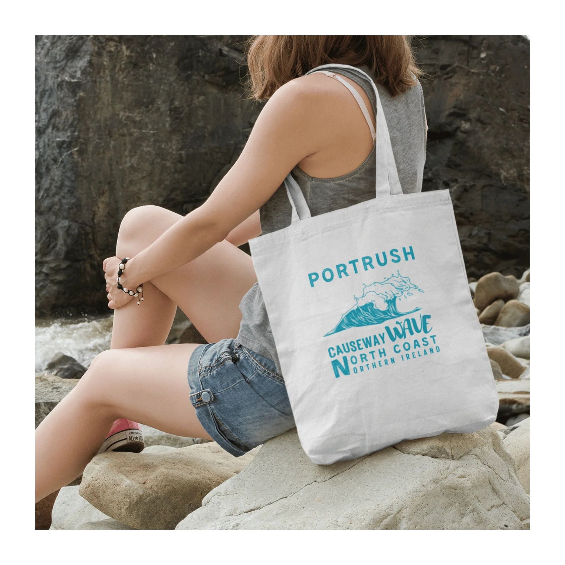 Person sitting on rocks by the sea with a tote bag featuring Portrush Causeway Wave North Coast Northern Ireland branding.