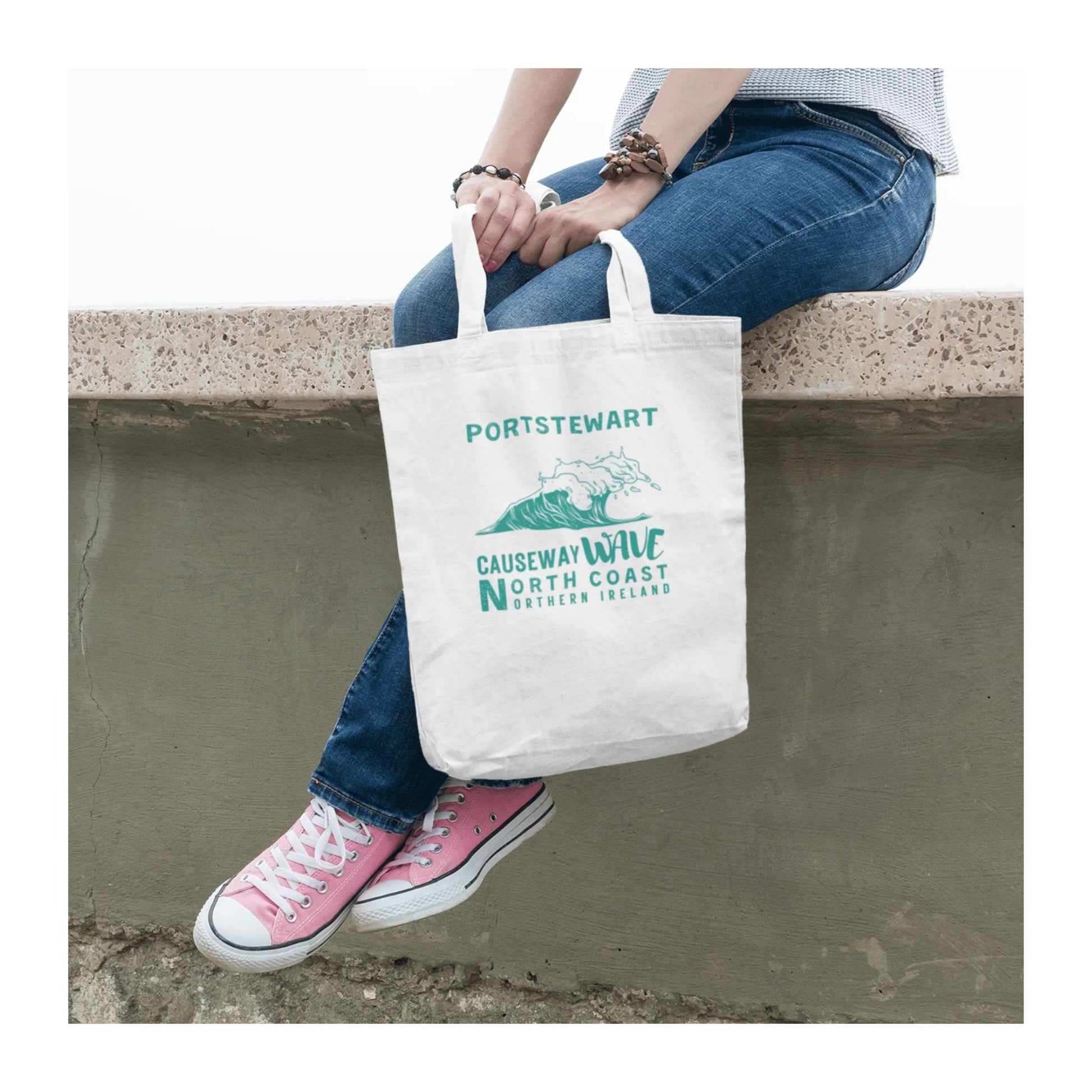 Person sitting on a ledge holding a tote bag with 'Portstewart Causeway Wave North Coast Northern Ireland' text and graphics.