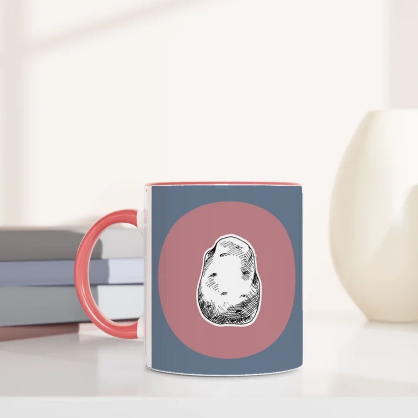Jane Austen inspired mug with a design of a potato on a pink circle against a blue background, placed on a surface with books and a vase.