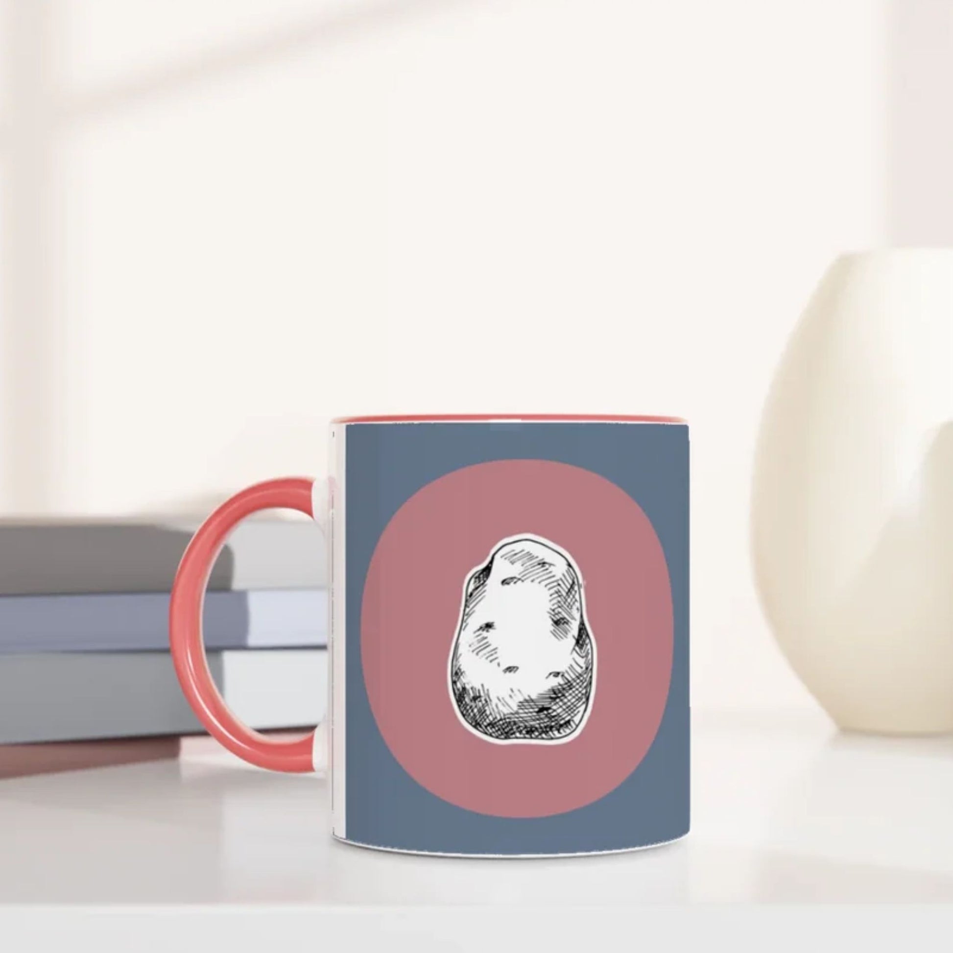 Jane Austen inspired mug with a design of a potato on a pink circle against a blue background, placed on a surface with books and a vase.